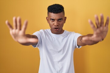 Young hispanic man standing over yellow background doing stop gesture with hands palms, angry and frustration expression