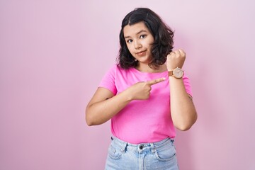 Young hispanic woman standing over pink background in hurry pointing to watch time, impatience, looking at the camera with relaxed expression