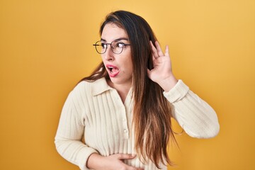 Young hispanic woman standing over yellow background smiling with hand over ear listening an hearing to rumor or gossip. deafness concept.