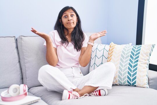 Young hispanic woman sitting on the sofa at home clueless and confused expression with arms and hands raised. doubt concept.