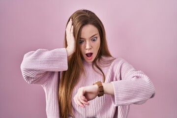 Young caucasian woman standing over pink background looking at the watch time worried, afraid of...