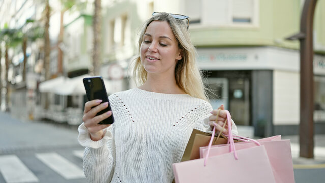 Young blonde woman going shopping holding bags using smartphone at street