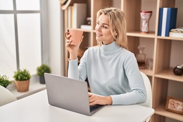 Young blonde woman using laptop and drinking coffee sitting on table at home