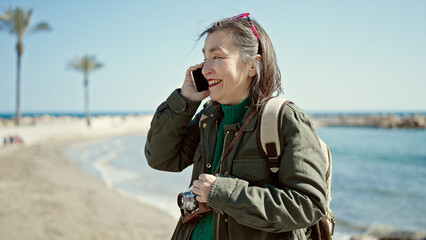 Mature hispanic woman with grey hair tourist wearing backpack speaking on the phone at seaside