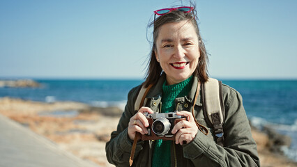 Mature hispanic woman with grey hair tourist wearing backpack holding vintage camera at seaside