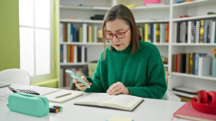 Mature hispanic woman with grey hair student using smartphone reading a book at library