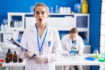 Blonde woman working at scientist laboratory scared and amazed with open mouth for surprise, disbelief face