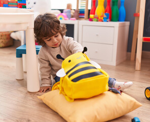 Adorable hispanic boy sitting on floor holding something of backpack at kindergarten