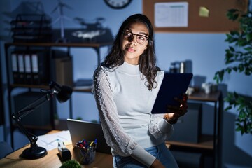 Young brazilian woman using touchpad at night working at the office smiling looking to the side and...