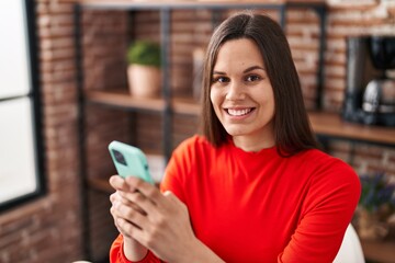 Young beautiful hispanic woman using smartphone sitting on table at home