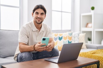 Fototapeta premium Young hispanic man using laptop and smartphone sitting on sofa at home