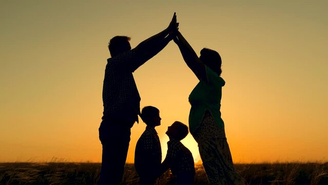 Taking care of children by parents. Happy family dreams of their own home in park in summer in sunshine. Children mom and dad are playing together building house with their hands, silhouette at sunset