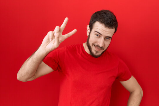 Young hispanic man wearing casual red t shirt smiling looking to the camera showing fingers doing victory sign. number two.