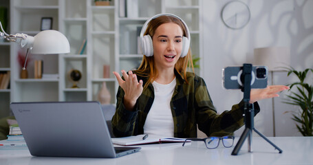 Woman influencer recording video blog on smartphone sitting at an home office desk. Girl sits front mobile phone camera on tripod records educational tutorial video recounts personal lifestyle blog