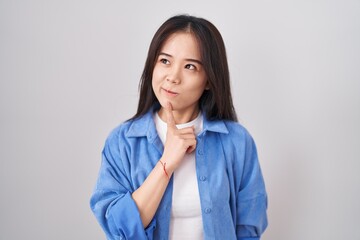 Young chinese woman standing over white background thinking concentrated about doubt with finger on chin and looking up wondering