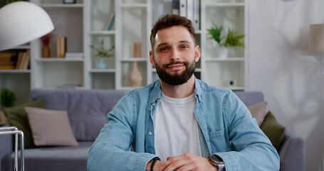 Smiling young adult man looking camera during webinar, online class, virtual meeting. Caucasian male person posing while sitting on living room at home.
