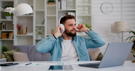 Handsome young man wearing headphones listening on smartphone favorite energetic music and dancing at home. Freelancer man relaxing, taking a break.