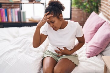 African american woman suffering for stomach and head ache sitting on bed at bedroom