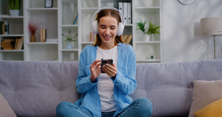 Young woman using phone to browse social media with headphones while relaxing on a sofa at home. Female resting stress relief with chilling happy peaceful face sits in living room.
