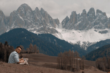 Naklejka na ściany i meble A man sits on a hill in front of a mountain with the mountains in the background. Hiking in the mountains. The alps.