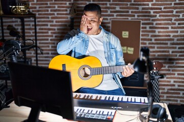 Hispanic young man playing classic guitar at music studio covering one eye with hand, confident smile on face and surprise emotion.
