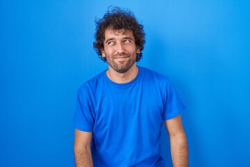 Fototapeta na wymiar Hispanic young man standing over blue background smiling looking to the side and staring away thinking.