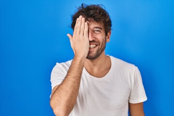 Hispanic young man standing over blue background covering one eye with hand, confident smile on face and surprise emotion.