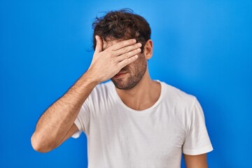 Hispanic young man standing over blue background covering eyes with hand, looking serious and sad. sightless, hiding and rejection concept