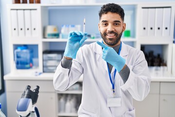 Hispanic man with beard working at scientist laboratory holding syringe smiling happy pointing with...