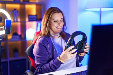 Young beautiful plus size woman streamer smiling confident holding headphones at gaming room