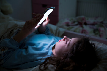 A child using smart phone lying in bed late at night, playing games, watching videos online, scrolling screen. Children's screen addiction and parent control concept. Child's room at night.  - obrazy, fototapety, plakaty