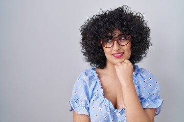 Young brunette woman with curly hair wearing glasses over isolated background looking confident at the camera smiling with crossed arms and hand raised on chin. thinking positive.