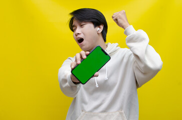 Asian man in beige hoodie using earphones, raising fist and showing his screen phone with green screen template best for advertisment. man showing mock up of phone.