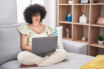 Fototapeta na wymiar Young brunette woman with curly hair using laptop sitting on the sofa at home relaxed with serious expression on face. simple and natural looking at the camera.