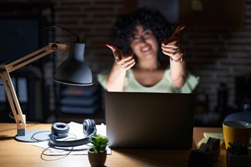 Young brunette woman with curly hair working at the office at night pointing fingers to camera with happy and funny face. good energy and vibes.