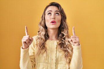 Obraz premium Young caucasian woman standing over yellow background amazed and surprised looking up and pointing with fingers and raised arms.