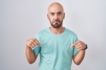 Middle age bald man standing over white background pointing down looking sad and upset, indicating...