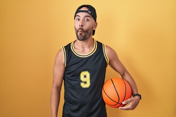Middle age bald man holding basketball ball over yellow background making fish face with lips, crazy and comical gesture. funny expression.