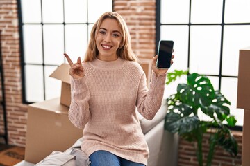 Hispanic woman moving to a new home showing smartphone screen smiling happy pointing with hand and finger to the side