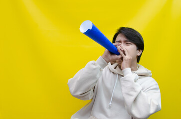 An ecstatic young Asian man in a cream hoodie whispering and shouting through a paper megaphone giving an announcement. asian man dancing and singing happily using paper as a megaphone.
