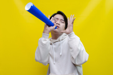 An ecstatic young Asian man in a cream hoodie whispering and shouting through a paper megaphone...