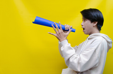 An ecstatic young Asian man in a cream hoodie whispering and shouting through a paper megaphone...