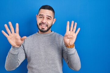 Hispanic man standing over blue background showing and pointing up with fingers number nine while...