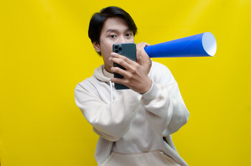 expressive Asian man holding phone while shouting to paper megaphone to give an offering and...