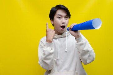 An ecstatic young Asian man in a cream hoodie whispering and shouting through a paper megaphone giving an announcement. asian man dancing and singing happily using paper as a megaphone.
