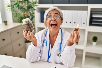 Middle age woman doctor holding cbd oil angry and mad screaming frustrated and furious, shouting...
