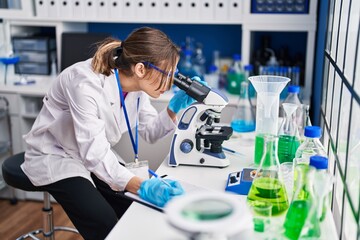 Fototapeta premium Young beautiful hispanic woman scientist using microscope writing on document at laboratory