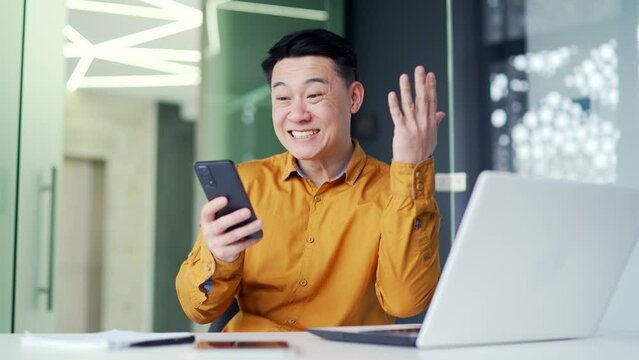 Smiling excited asian businessman celebrating success by reading good news on smartphone while sitting at desk at workplace in modern office. Happy employee shocked, surprised by a pleasant message