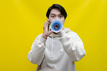 expressive Asian man holding phone while shouting to paper megaphone to give an offering and...