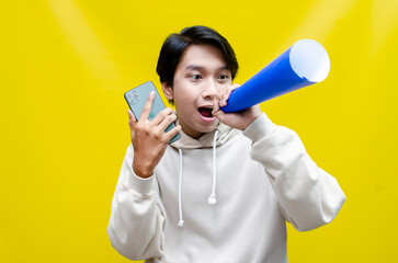 expressive Asian man holding phone while shouting to paper megaphone to give an offering and...